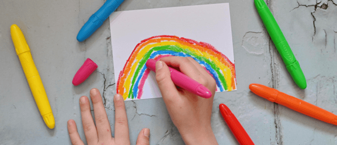 A child's hands use crayons to draw a rainbow