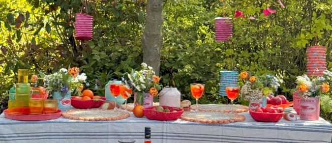 A long table underneath a tree in a garden is laid with colourful placemats, lanterns and baskets