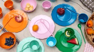 Bright melamine plates, bowls, cups, and cutlery arranged neatly on a patterned tablecloth, Ideal for picnic.