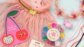 A flat lay of colorful children’s accessories on a light blue background, including a pink tulle dress with a gold butterfly belt, a cherry-shaped card labeled ‘Stick-on earrings,’ a pack of pink hair clips, a circular bead set labeled ‘Make your own jewelry set,’ a pink unicorn figurine, and a headband decorated with pink flowers and multicoloured ribbons.