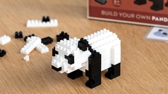A small black-and-white building block panda figure, partially assembled, sitting on a wooden table next to its packaging box labelled 'Build Your Own Panda.