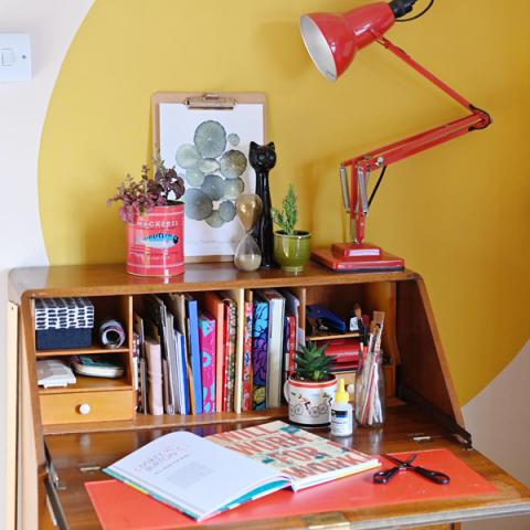 A desk filled with books and notepads, with a plant in a red tin and a red desk lamp