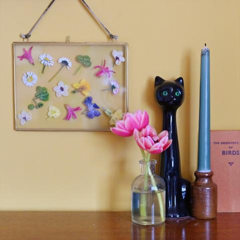 Flower art in a brass frame next to a candle and vase, against a yellow wall