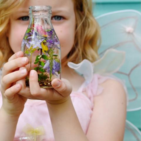 A young girl wearing fairy wings holds a glass jar filled with flowers