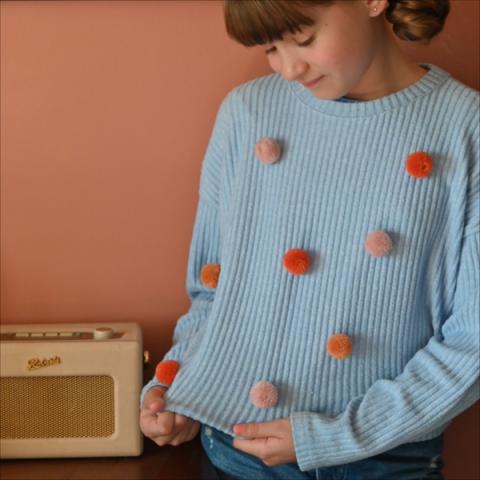 A girl wears a light blue jumper decorated with pink pompoms