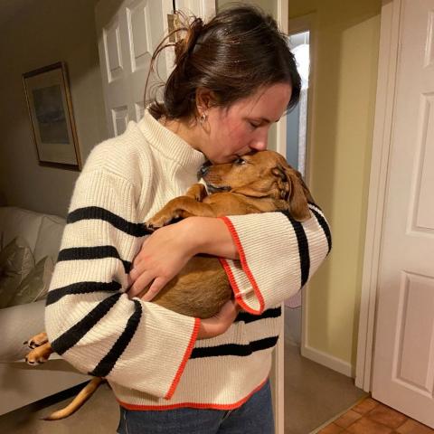 A woman holds a sausage dog and is kissing her on the head