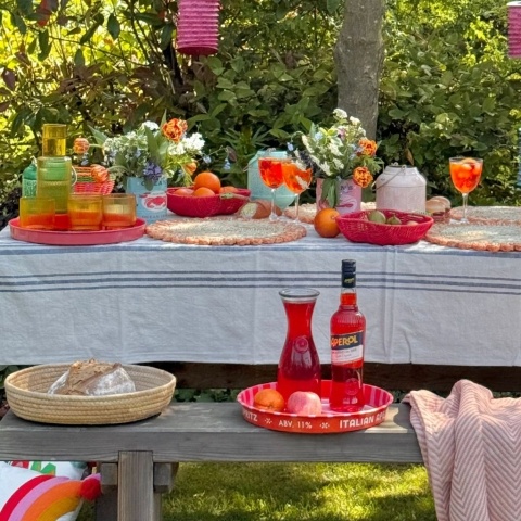 An outdoor table is laid with drinks and flowers, including a carafe and serving tray on a bench