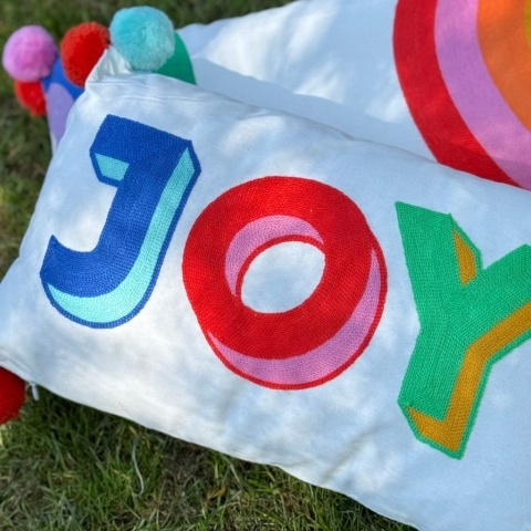 A white cushion with the word 'JOY' in colourful letters lays on a lawn
