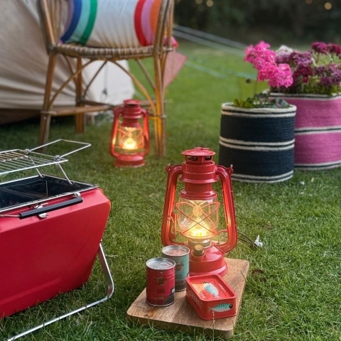 A red hurricane lantern sits on a wooden board on the grass, next to a portable BBQ