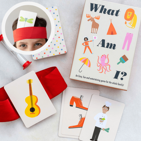 Children's "What Am I?" guessing game with illustrated cards, a red headband, and a sand timer arranged on a tabletop.