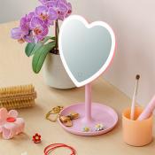 Pink heart-shaped light-up vanity mirror on a wooden surface, next to a potted orchid plant, a double wooden brush, and jewelry.