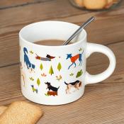 A white ceramic mug - Dog park, with a metal spoon inside. The mug is decorated with colourful illustrations of dogs and small trees.
