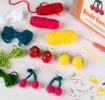 Crochet kit packaging placed beside three finished crocheted earrings shaped like strawberries, lemons, and cherries