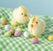 Two fluffy chicks on a green gingham cloth surrounded by small coloured eggs