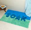 A blue tufted bath mat with the word “SOAK” in white displayed on a bathroom floor.