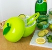 Apple-shaped ice bucket with lid and tongs, surrounded by citrus and drinks on a table.