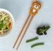 Training chopsticks with a bear topper beside a bowl of noodles and vegetables.