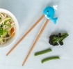 Training chopsticks with a whale topper beside a bowl of noodles and vegetables.