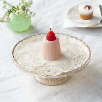 Pink jelly-shaped candle topped with a decorative red strawberry displayed on a decorative cake stand.