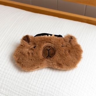 Light brown plush capybara‑shaped travel eye mask with stitched facial details
