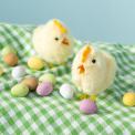 Two fluffy chicks on a green gingham cloth surrounded by small coloured eggs