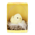 A fluffy yellow chick decoration sitting in a small wicker basket with an egg, displayed inside a yellow box.
