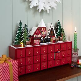 Red advent calendar with 24 gold-numbered drawers and a snowy village scene on top