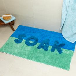A blue tufted bath mat with the word “SOAK” in white displayed on a bathroom floor.