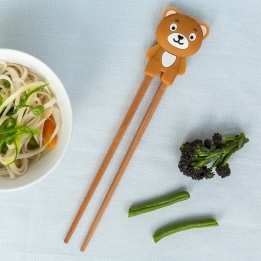 Training chopsticks with a bear topper beside a bowl of noodles and vegetables.