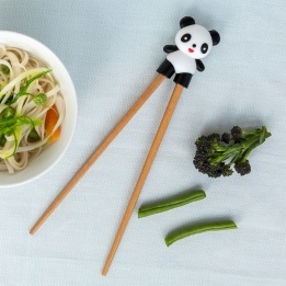 Training chopsticks with a panda topper beside a bowl of noodles and vegetables.