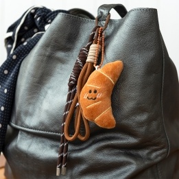 Plush croissant bag charm with brown cord strap and metal clip attachment