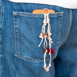 Beige cord phone strap with bead and flower charms shown hanging from a phone in a denim pocket.