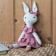 Knitted rabbit toy with long ears, wearing a pink dress with a ruffled collar and a matching bow on one ear