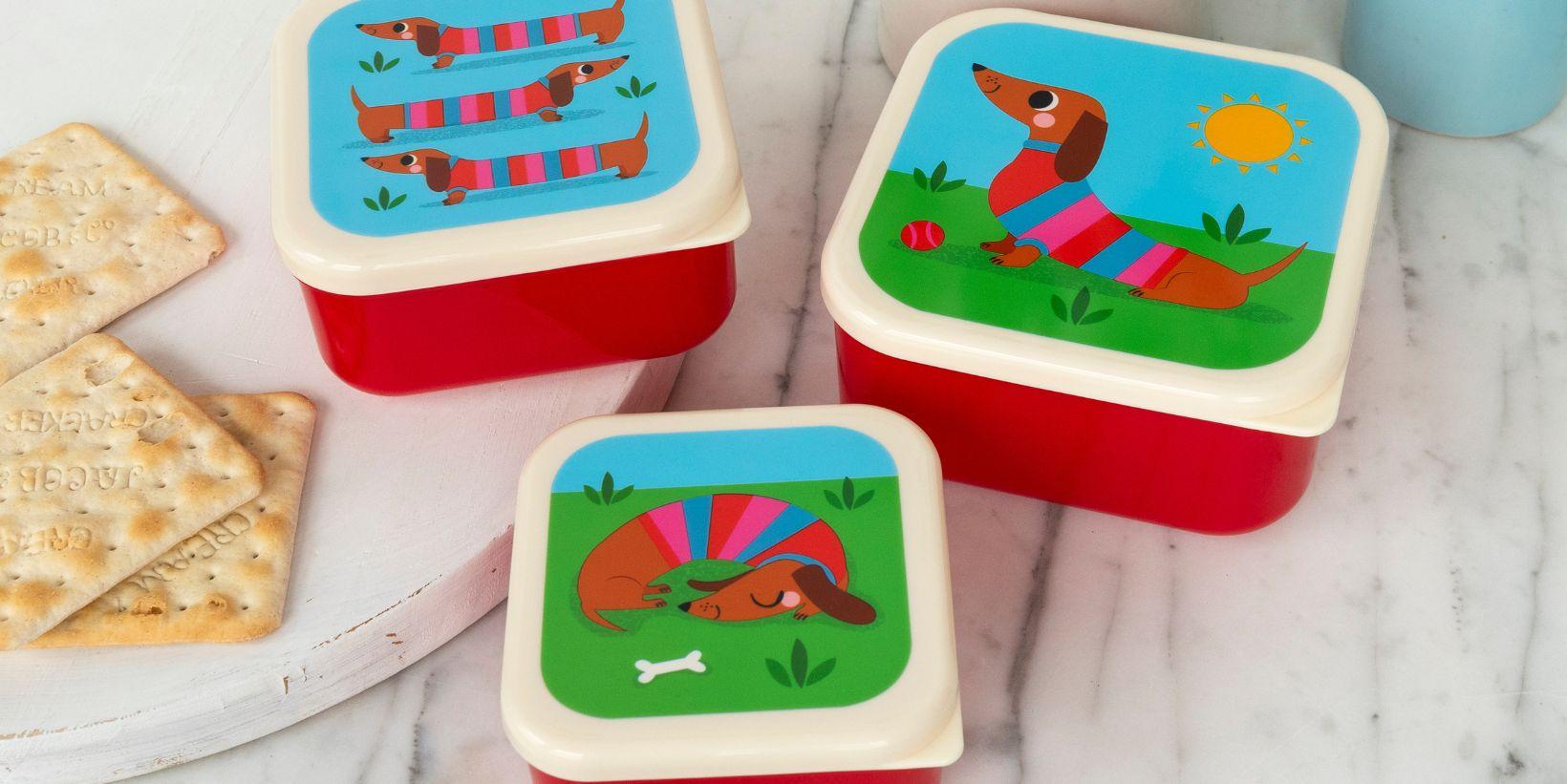 Three snack boxes of different sizes with a sausage dog design sit on a table next to some crackers