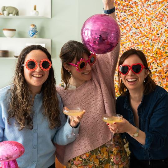 Three women wearing flower-shaped sunglasses hold pink disco balls and cocktail glasses