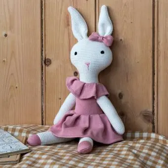 Knitted rabbit toy with long ears, wearing a pink dress with a ruffled collar and a matching bow on one ear