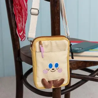 Beige and yellow crossbody bag with a front pocket featuring a smiling face design and two small brown plush feet at the bottom.