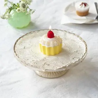 Cupcake‑shaped candle with white frosting and a strawberry topper displayed on a decorative cake stand
