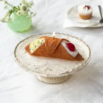Brown pastry-shaped candle with cream and red cherry decorations displayed on a cake stand.