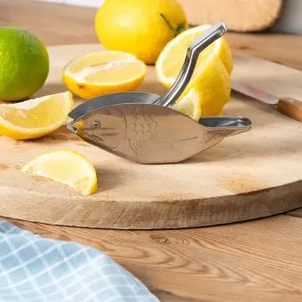 Stainless steel fish‑shaped lemon squeezer on a wooden board with cut lemons.