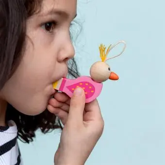 Child blowing a bird‑shaped wooden whistle with a pink body, wooden head, and orange beak.