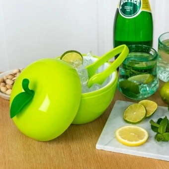 Apple-shaped ice bucket with lid and tongs, surrounded by citrus and drinks on a table.