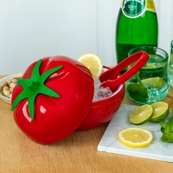 Tomato-shaped ice bucket with lid and tongs, surrounded by citrus and drinks on a table.