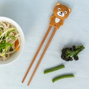 Training chopsticks with a bear topper beside a bowl of noodles and vegetables.
