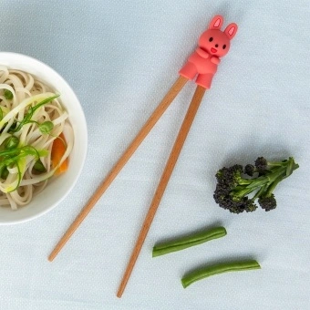 Training chopsticks with a bunny topper beside a bowl of noodles and vegetables.