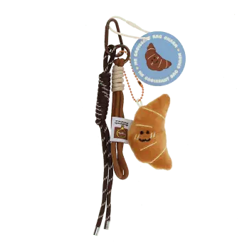 Plush croissant bag charm with brown cord strap and metal clip attachment with tag.
